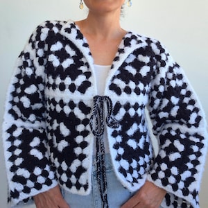 May include: A black and white crochet cardigan featuring a granny square pattern with long sleeves and a front tie closure made of braided yarn. The garment is constructed from fuzzy, textured yarn in a repeating block design.