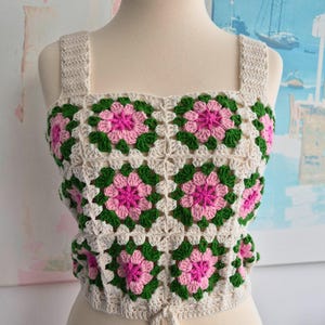 May include: A sleeveless crochet crop top featuring a square neckline and wide straps. The garment is constructed from joined granny squares in a floral pattern, showcasing cream, forest green, and bright pink yarn with a tie closure at the hem.
