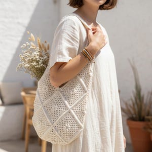 May include: Cream-colored, hand-crocheted tote bag with a geometric diamond pattern. The bag is filled with dried flowers and carried over the shoulder. The person is wearing a cream-colored dress and wooden beaded bracelets.