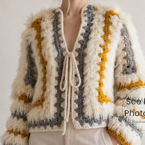 Puede incluir: Chaqueta de crochet color crema, con frente abierto y textura peluda. La chaqueta presenta rayas verticales grises y amarillo mostaza, con cierre de lazo en el escote. Las mangas son largas y acampanadas, y la chaqueta está hecha a mano.