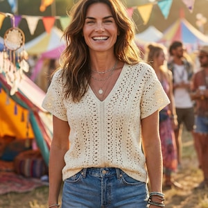 Puede incluir: Una mujer con un top de crochet crema con cuello en V y vaqueros azules. Sonr&iacute;e frente a una carpa colorida y atrapasue&ntilde;os, lo que sugiere un festival o evento al aire libre.