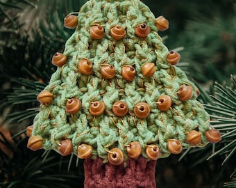 May include: A triangular crochet Christmas tree ornament in light green yarn with metallic gold thread accents. The tree is decorated with small, round, brown wooden beads arranged in horizontal rows and features a solid dark brown rectangular trunk.