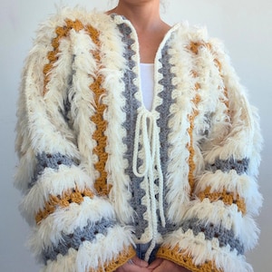 May include: A crochet cardigan featuring a granny square pattern in mustard yellow, gray, and cream. The garment is accented with long, shaggy cream-colored fringe throughout the body and sleeves, and includes a tie closure at the center front.