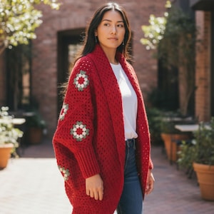 May include: A red, open-front crochet cardigan with floral accents on the sleeves. The cardigan is worn over a white top and blue jeans. The sleeves have a pattern of green, white, and red floral designs. The cardigan is a cozy, handmade garment.