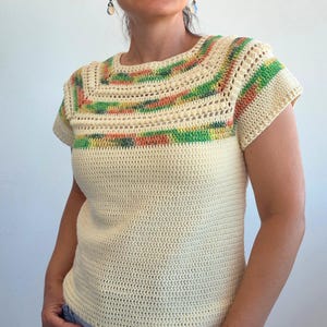 May include: A short-sleeved crochet top in cream yarn featuring a square neckline with horizontal bands of variegated green, orange, and yellow yarn. The garment is constructed with a dense stitch pattern on the body and a textured stitch detail.