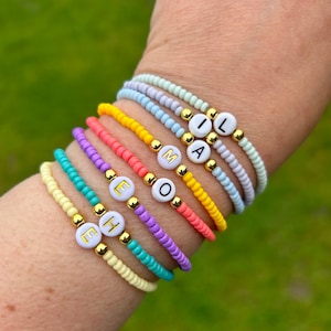 May include: A set of colourful beaded bracelets with white letter beads spelling out the word "EMMA" in all lowercase letters. The bracelets are stacked on a person's wrist.