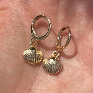 May include: A pair of gold-coloured hoop earrings with seashell charms. The earrings have a hinged closure and a small, detailed seashell pendant. The earrings are shown against a blurred background.