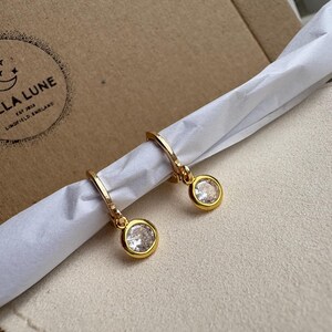 May include: Gold-coloured hoop earrings, each with a clear, round gemstone pendant. The earrings are presented on white tissue paper, with a brown cardboard box in the background. The Bella Lune logo is visible on the box.