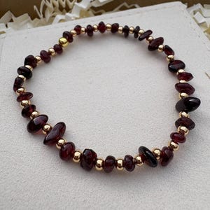 May include: A bracelet with dark red gemstones and small gold-coloured beads. The gemstones are irregularly shaped and interspersed with the gold beads. The bracelet is circular, with the beads strung on a flexible cord. The gemstones vary in size.