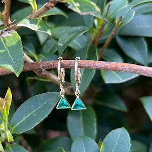 May include: Gold-coloured hoop earrings with triangular, emerald-green pendants. The earrings hang from a brown branch, set against a backdrop of green leaves and foliage. The earrings are a simple, elegant design, suitable for everyday wear.