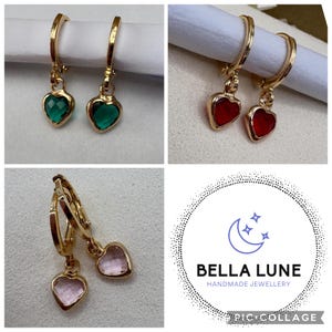 May include: Gold-toned hoop earrings featuring dangling heart-shaped charms in green, red, and light pink. Each charm is faceted and set in a gold-coloured metal frame. The image includes a logo with a crescent moon and the text BELLA LUNE.
