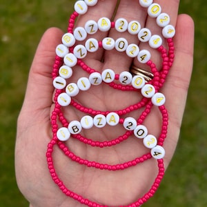 May include: Five pink beaded bracelets with white beads that spell out "IBIZA 2024" in gold letters.