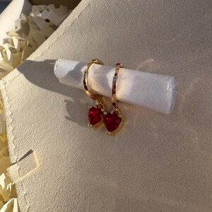 May include: Gold-coloured hoop earrings, each with a red heart-shaped gemstone. The earrings are presented on a rolled white paper, against a neutral backdrop. The earrings are in a box with packing material.
