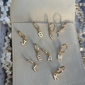 May include: Gold-tone hoop earrings with white enamel letter charms spelling out "TEAM" and other letters. The earrings are presented on a light grey surface, with a soft, neutral background.
