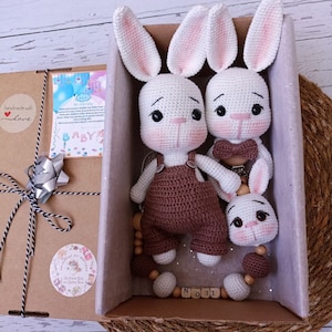 Könnte beinhalten: Eine Geschenkbox mit zwei weißen Häkel-Hasenspielzeugen, einem Holzperlen-Beißring und einem Holz-Namensschild mit der Aufschrift "LEON". Die Box ist mit einem Etikett verziert, auf dem "Handgefertigt mit Liebe" steht, und einem silbernen Band.