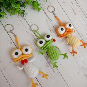 May include: Three handmade crochet keychains in the shape of whimsical birds and a frog. The keychains feature large googly eyes, beaks, and feet in yellow, orange, white, and green. Each keychain has a silver metal ring.