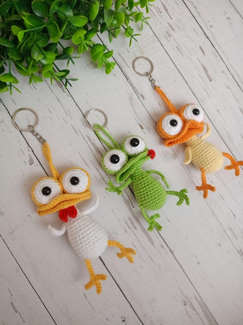 May include: Three handmade crochet keychains in the shape of whimsical birds and a frog. The keychains feature large googly eyes, beaks, and feet in yellow, orange, white, and green. Each keychain has a silver metal ring.
