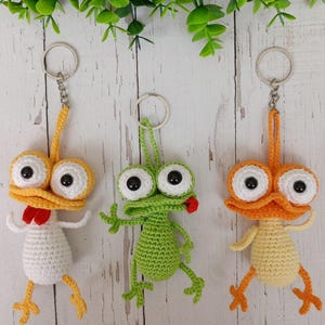 May include: Three handmade crochet keychains featuring whimsical cartoon characters. One is white with a red accent, another is green, and the third is orange. Each has large, googly eyes and a metal keyring.