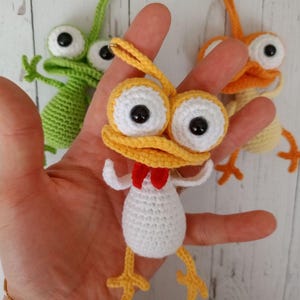 May include: Handmade crochet keychains in the shape of whimsical creatures. The central keychain is yellow, white, and red, with large white-rimmed eyes and a red bow tie. Other keychains are green and orange. The keychains are held in a hand.