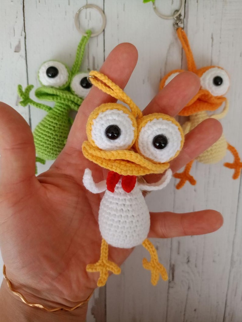 May include: Handmade crochet keychains in the shape of whimsical creatures. The central keychain is yellow, white, and red, with large white-rimmed eyes and a red bow tie. Other keychains are green and orange. The keychains are held in a hand.