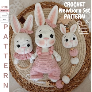 Può includere: Modello all'uncinetto per un set neonato con tre giocattoli amigurumi a forma di coniglio rosa e bianco. Il coniglio più grande indossa una salopette rosa e ha un anello di legno attaccato al braccio. Il coniglio più piccolo è attaccato a un giocattolo da dentizione con perline di legno con il nome "Emilia" scritto sulle perline. Il modello è disponibile in formato PDF.