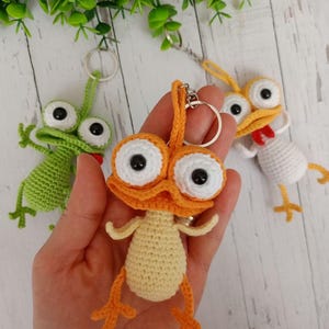 May include: Crocheted keychains in the shape of cartoon creatures. The central keychain is orange and yellow, with large white-rimmed eyes and black pupils. The other keychains are green and white. The keychains are held in a hand.