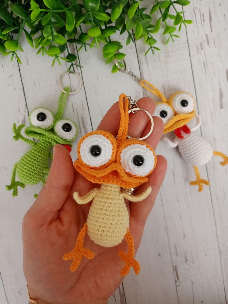 May include: Crocheted keychains in the shape of cartoon creatures. The central keychain is orange and yellow, with large white-rimmed eyes and black pupils. The other keychains are green and white. The keychains are held in a hand.