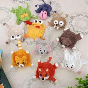 Puede incluir: Una colección de diez marcapáginas de ganchillo planos y coloridos con varias criaturas, incluyendo una rana verde, un burro morado, un pájaro amarillo, un oso marrón, un elefante gris, un conejo blanco y un zorro naranja con detalles de hilo.