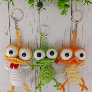 May include: Three handmade crochet keychains featuring whimsical creature designs. One is yellow and white with a red bow tie, another is green, and the third is orange and yellow. Each has large eyes and a metal keyring.