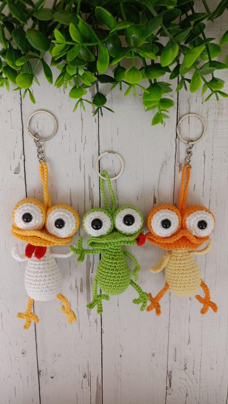 May include: Three handmade crochet keychains featuring whimsical creature designs. One is yellow and white with a red bow tie, another is green, and the third is orange and yellow. Each has large eyes and a metal keyring.
