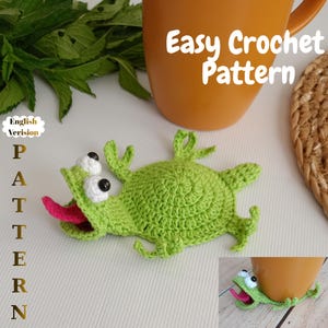 Könnte beinhalten: Ein grüner gehäkelter Frosch-Untersetzer mit weißen Augen und einer rosa Zunge. Der Untersetzer befindet sich neben einer braunen Tasse. Der Text auf dem Bild lautet "Easy Crochet Pattern" und "English Version Pattern".