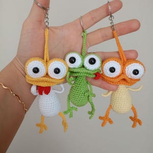 May include: Three handmade crochet keychains in yellow, green, and orange. Each keychain features a whimsical creature with large, googly eyes, small arms, and legs. The yellow creature has a red bow tie, the green one has a red mouth, and the orange one has a yellow body.