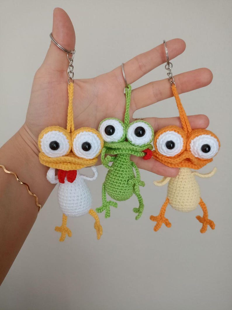 May include: Three handmade crochet keychains in yellow, green, and orange. Each keychain features a whimsical creature with large, googly eyes, small arms, and legs. The yellow creature has a red bow tie, the green one has a red mouth, and the orange one has a yellow body.