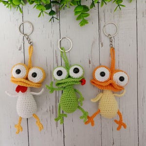 May include: Three handmade crochet keychains. One is a white chicken with yellow feet and a red comb. The second is a green frog with a red tongue. The third is an orange duck with yellow feet. All have large googly eyes.