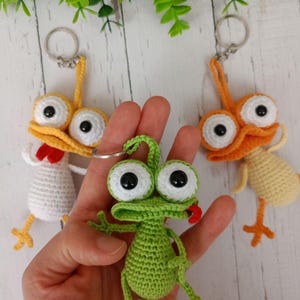May include: Crocheted keychains featuring whimsical frog and chicken designs. The frog is green with a red tongue, and the chickens are yellow and white. Each keychain has large, googly eyes and a metal ring.