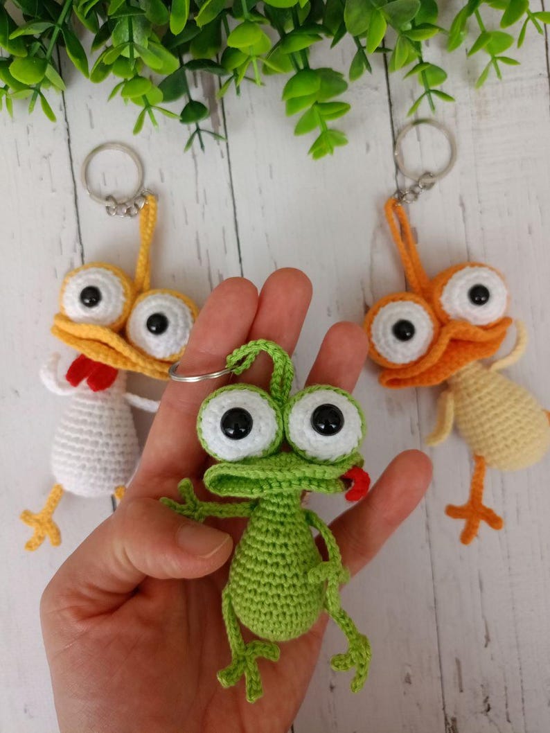 May include: Crocheted keychains featuring whimsical frog and chicken designs. The frog is green with a red tongue, and the chickens are yellow and white. Each keychain has large, googly eyes and a metal ring.