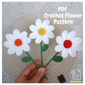 Puede incluir: Patrón de crochet PDF para tres flores de margarita blancas con centros de diferentes colores: naranja, amarillo y rojo. Las flores están tejidas a crochet y tienen tallos y hojas verdes.
