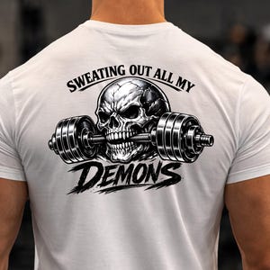 Może przedstawiać: Biała koszulka z czarno-białą grafiką czaszki trzymającej sztangę. Tekst "SWEATING OUT ALL MY DEMONS" jest wydrukowany nad i pod wzorem czaszki i sztangi. Koszulka jest wykonana z miękkiego materiału.