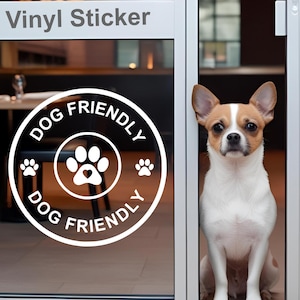 Könnte beinhalten: Weißer Vinyl-Aufkleber auf einer Glastür mit dem Text "Dog Friendly" in einem Kreis mit Pfotenabdrücken. Ein kleiner brauner und weißer Hund schaut durch die Tür.