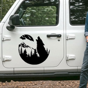 May include: Black silhouette decal of a howling wolf, mountains, and trees, with a crescent moon. The decal is on a white vehicle door. The design depicts a wilderness scene.