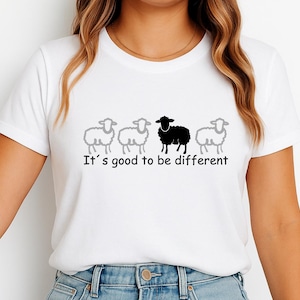 Op de afbeelding: Wit T-shirt met een afbeelding van vier schapen, waarvan één zwart en drie grijs. De tekst "It's good to be different" staat eronder in het zwart gedrukt. Eenvoudig ontwerp.