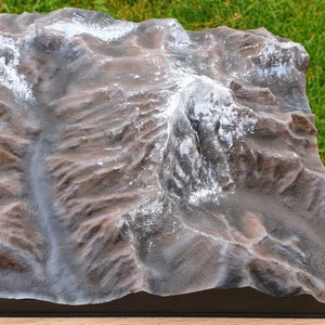 Puede incluir: Un modelo 3D detallado de una cordillera montañosa, con un pico cubierto de pintura blanca para representar la nieve. El modelo está hecho de un material marrón oscuro y se muestra sobre una base de madera.