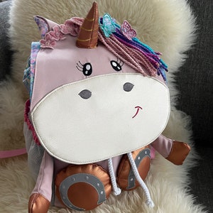 Könnte beinhalten: Ein rosa Rucksack in Form eines Einhorns mit einem goldenen Horn, rosa und violettem Garnhaar und Schmetterlingsverzierungen. Der Rucksack hat eine weiße Vorderseite mit gestickten Augen und einem Mund. Der Rucksack hat zwei kupferfarbene hufeisenförmige Füße.