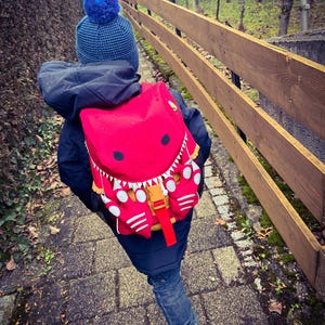 Op de afbeelding: Een kind met een marineblauwe jas, een spijkerbroek en een blauwe gebreide muts met een pompon loopt op een stenen pad. Een felrode rugzak met een cartoonmonsterontwerp zit op de rug van het kind. De rugzak heeft witte accenten en een gele gesp.