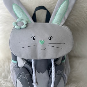 Könnte beinhalten: Ein grauer Rucksack in Hasenform mit grünen Ohren und einer weißen Blume. Der Rucksack hat einen weißen Kordelzug und graue Füße.