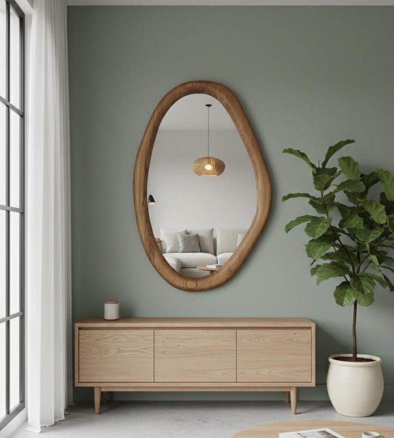 May include: An oval-shaped wooden mirror hangs above a light wood cabinet with three doors. A potted plant with large green leaves sits to the right. The mirror reflects a living room with a sofa and a pendant light.