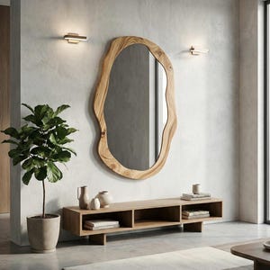 May include: A large, organic-shaped wooden mirror hangs on a textured white wall. Below, a low wooden console holds decorative vases and books. A potted plant and two wall sconces flank the mirror, creating a modern, minimalist aesthetic.
