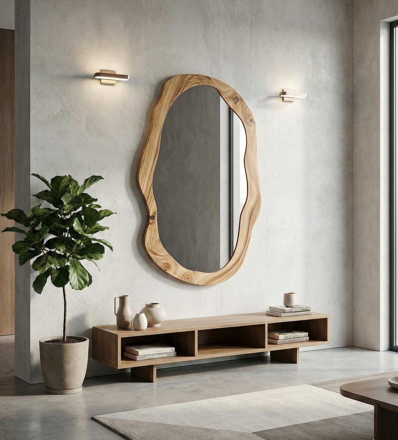 May include: A large, organic-shaped wooden mirror hangs on a textured white wall. Below, a low wooden console holds decorative vases and books. A potted plant and two wall sconces flank the mirror, creating a modern, minimalist aesthetic.
