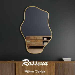 Puede incluir: Un espejo de forma abstracta con marco dorado sobre un fondo gris oscuro. El espejo refleja un armario de madera con estantes y libros. El texto "Rossena Mirror Design" est&aacute; debajo del espejo.