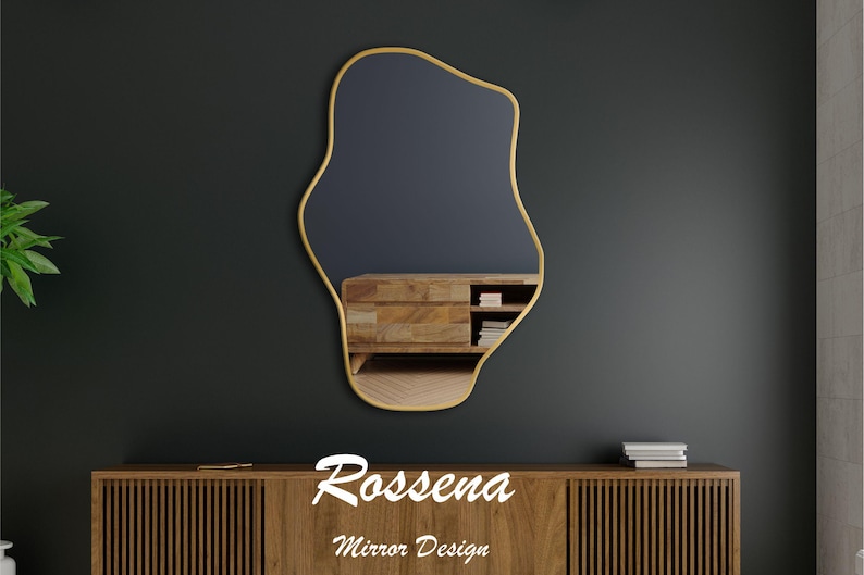 Puede incluir: Un espejo de forma abstracta con marco dorado sobre un fondo gris oscuro. El espejo refleja un armario de madera con estantes y libros. El texto "Rossena Mirror Design" est&aacute; debajo del espejo.